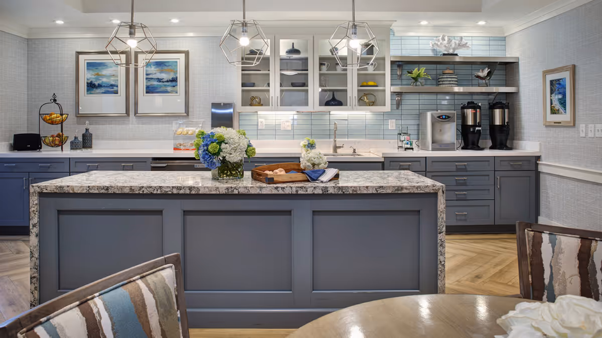 Modern kitchen area with a large marble island in the center, decorated with vases of blue and white flowers. The kitchen has gray cabinets, a light blue tiled backsplash, and open shelves with decorative items. Two geometric pendant lights hang above the island. There are framed paintings on the walls and a round table with patterned chairs in the foreground.