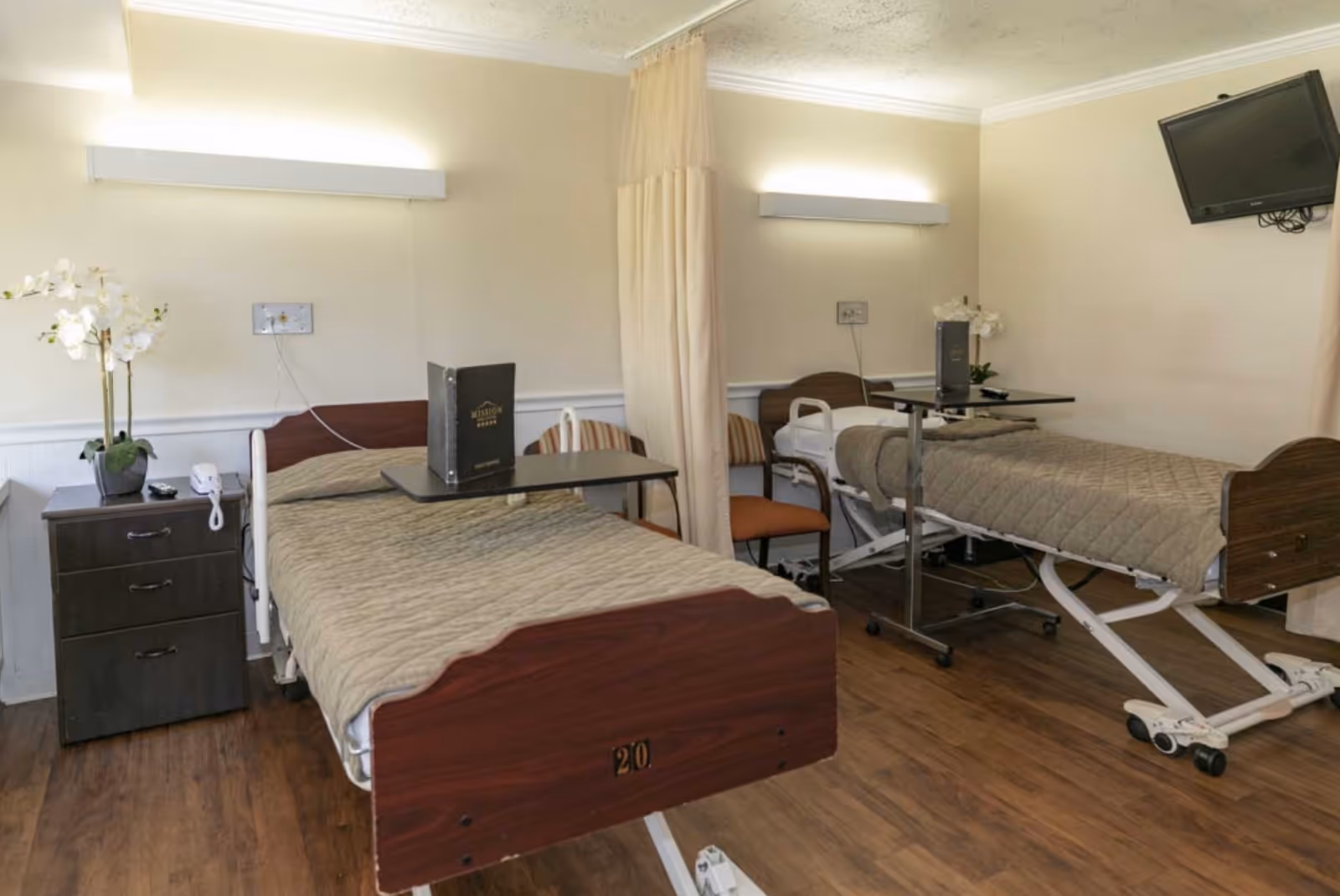 A room with two hospital-style beds separated by a privacy curtain. Each bed has a beige quilted blanket and an overbed table with a menu or brochure on it. There are chairs beside each bed, a nightstand with a telephone and a potted orchid, wall-mounted lights above each bed, and a wall-mounted television in the corner. The floor is wooden and the walls are light-colored.