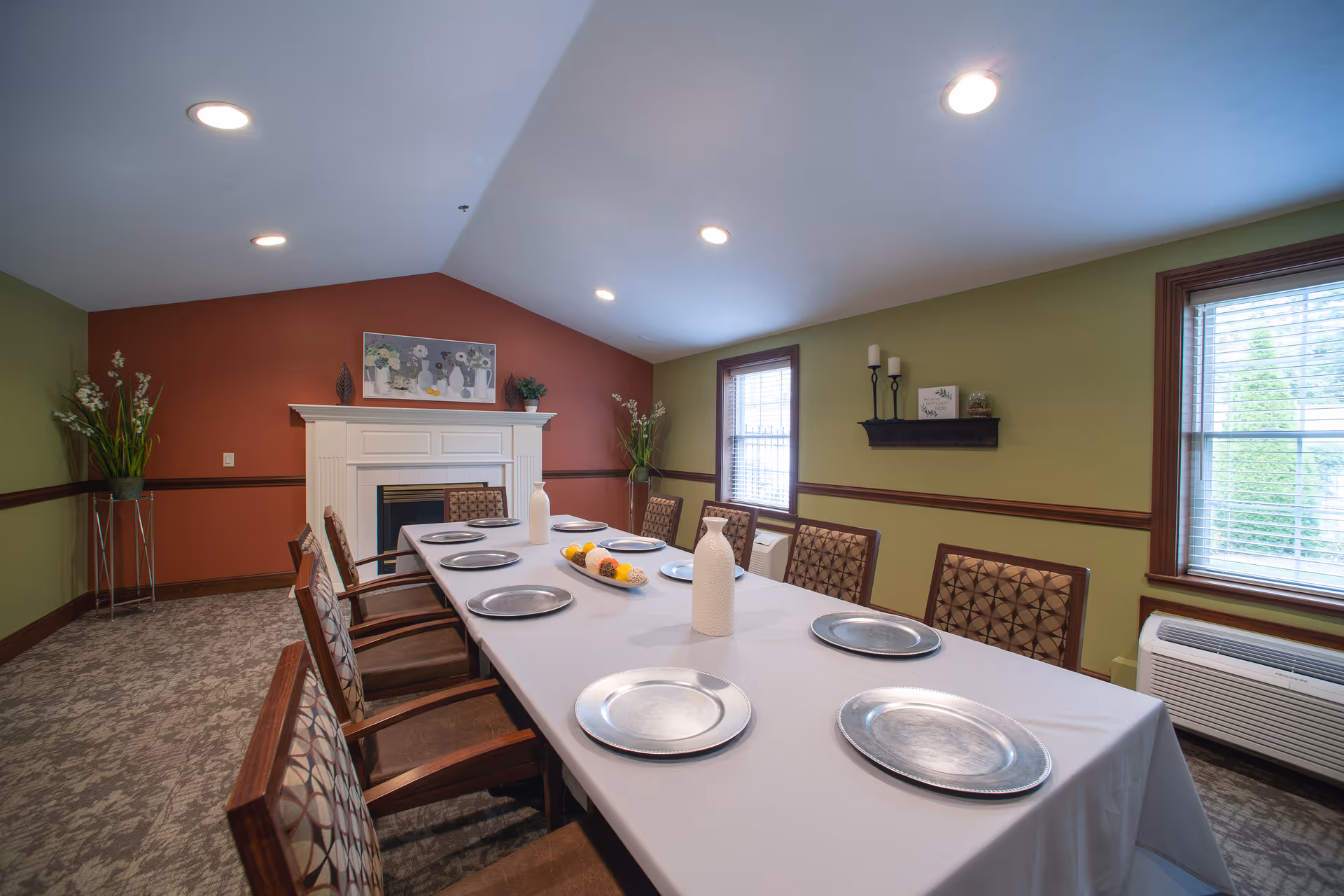 A dining room with a long table covered with a white tablecloth, set with silver plates and decorative white vases. The room has green and red walls, two windows with blinds, a white fireplace with a painting above it, and floral arrangements in the corners.