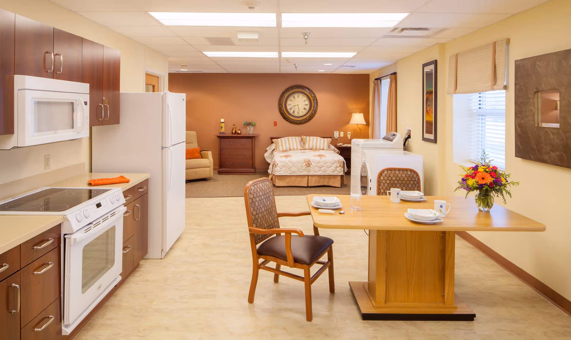 A bright and clean studio apartment in a senior living facility featuring a kitchen area with white appliances and wooden cabinets on the left, a wooden dining table set for two with a flower vase on the right, and a sleeping area with a bed, nightstand, lamp, and clock on the far wall. There is also a washing machine and dryer next to the bed, a comfortable armchair, and windows with blinds letting in natural light.