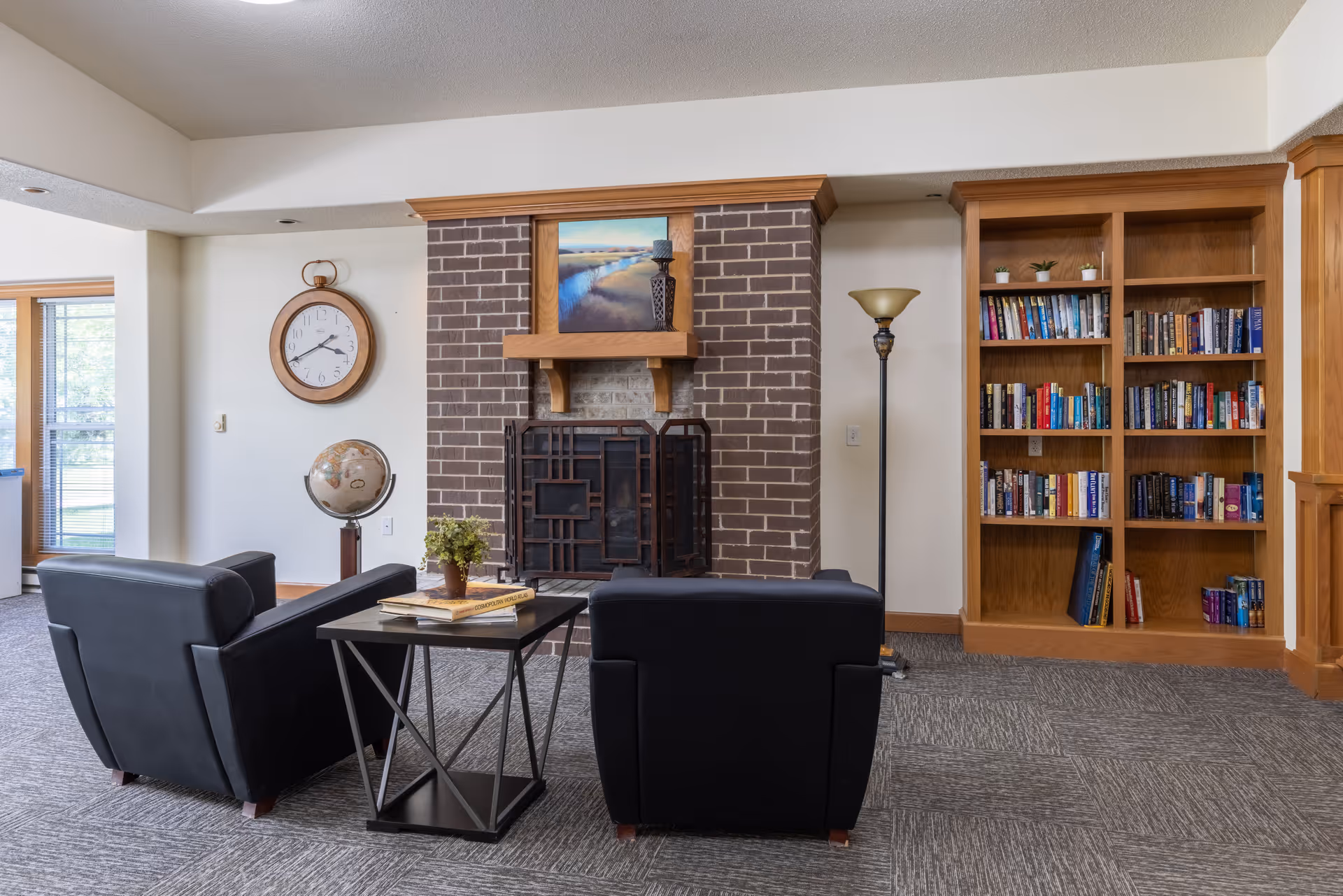 A cozy lounge with two black armchairs facing a brick fireplace, a side table, floor lamp, and a wooden bookshelf.