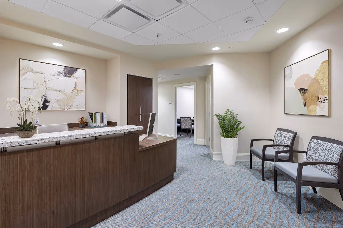 Reception area in a senior living facility with a wooden front desk topped with a granite counter, a computer monitor, and decorative items including a potted orchid and file organizers. The room has light-colored walls, abstract artwork on the walls, blue patterned carpet, two cushioned chairs with patterned backs, and a tall white planter with a green plant. A hallway with open doors is visible in the background.