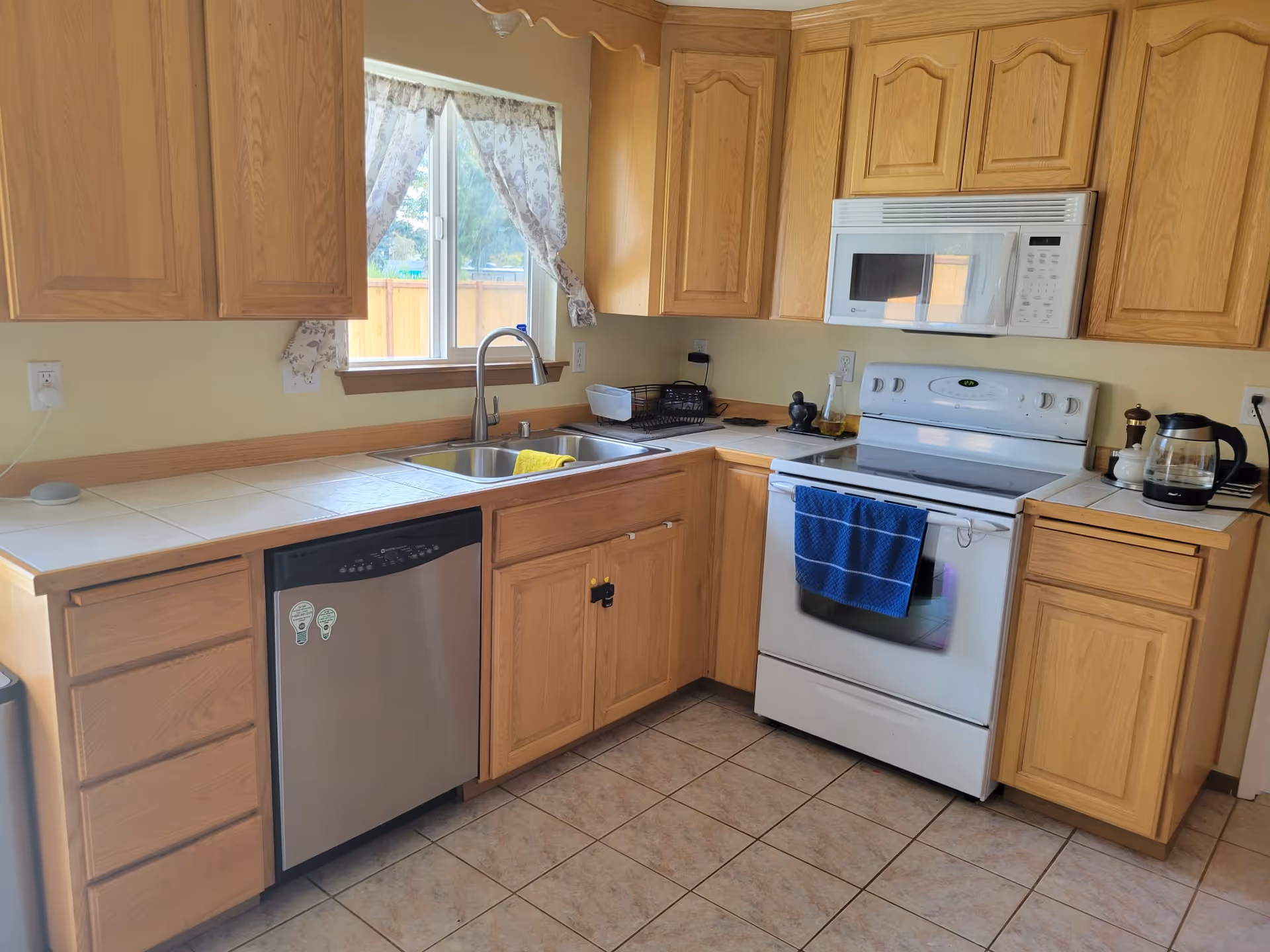 A kitchen with wooden cabinets, a white stove with a blue towel hanging on the handle, a white microwave above the stove, a stainless steel dishwasher, a double sink under a window with lace curtains, and a coffee maker on the counter.