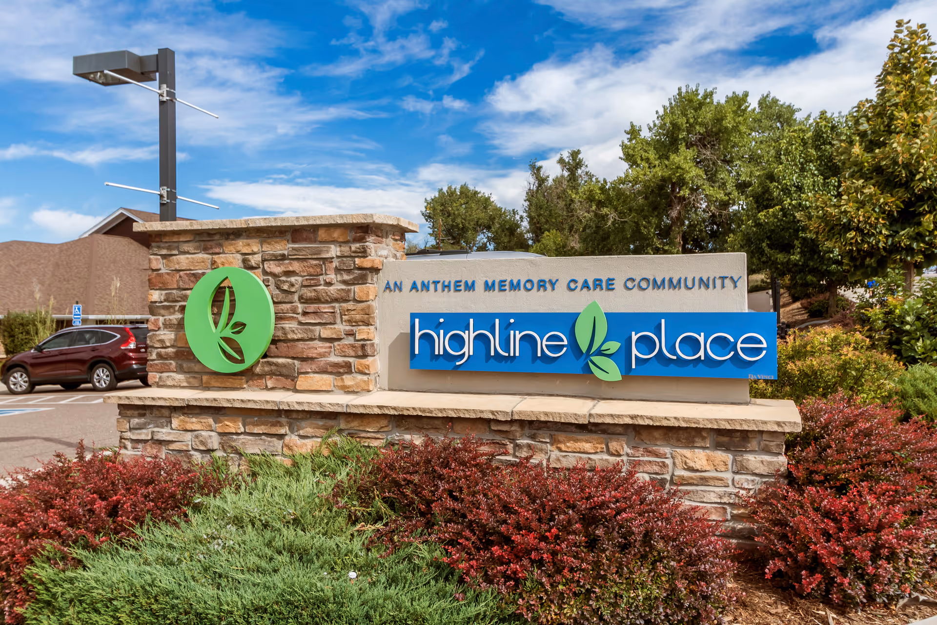 Stone and concrete sign for Highline Place, an Anthem Memory Care Community, surrounded by green and red shrubs with a blue sky and some trees in the background.