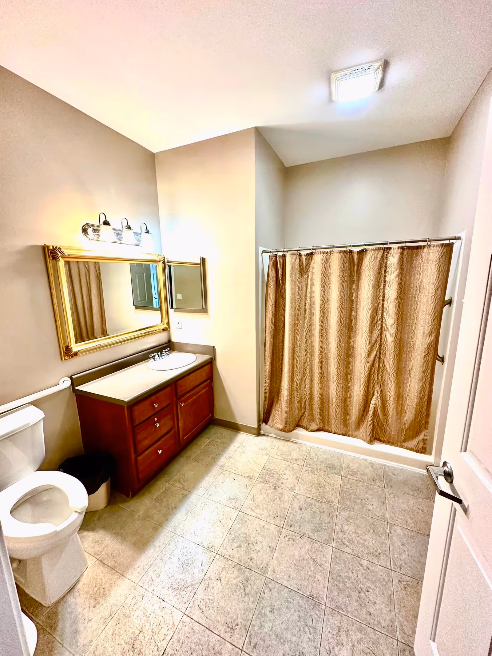 Well-lit bathroom with a vanity and mirror, toilet, and a shower with a beige curtain.