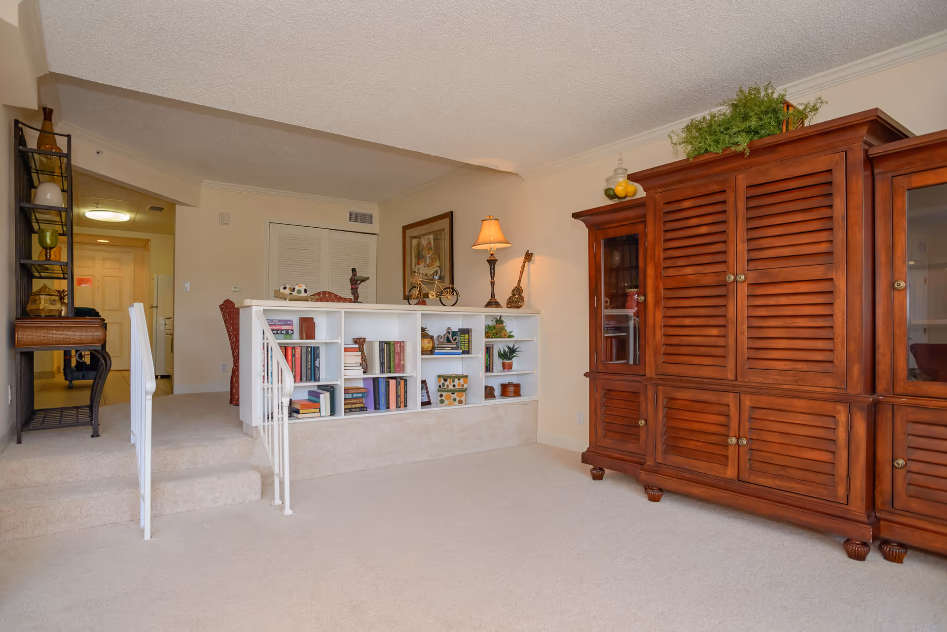 Interior of a senior living facility showing a carpeted living room area with a large wooden cabinet on the right, a white built-in bookshelf with various books and decorative items in the center, and a small set of carpeted stairs with white railings leading up to a hallway and kitchen area in the background.
