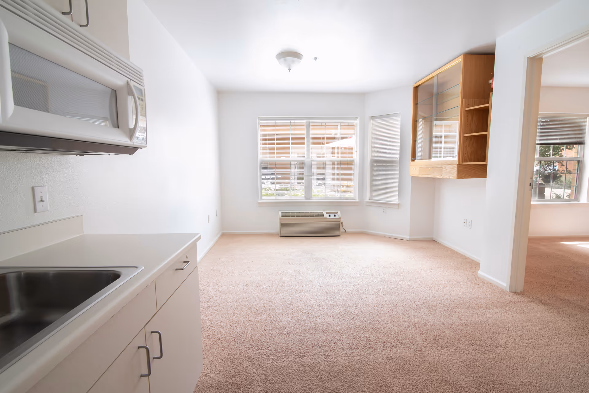 Bright empty apartment living room with a small kitchenette on the left, bay windows, and carpeted floor.