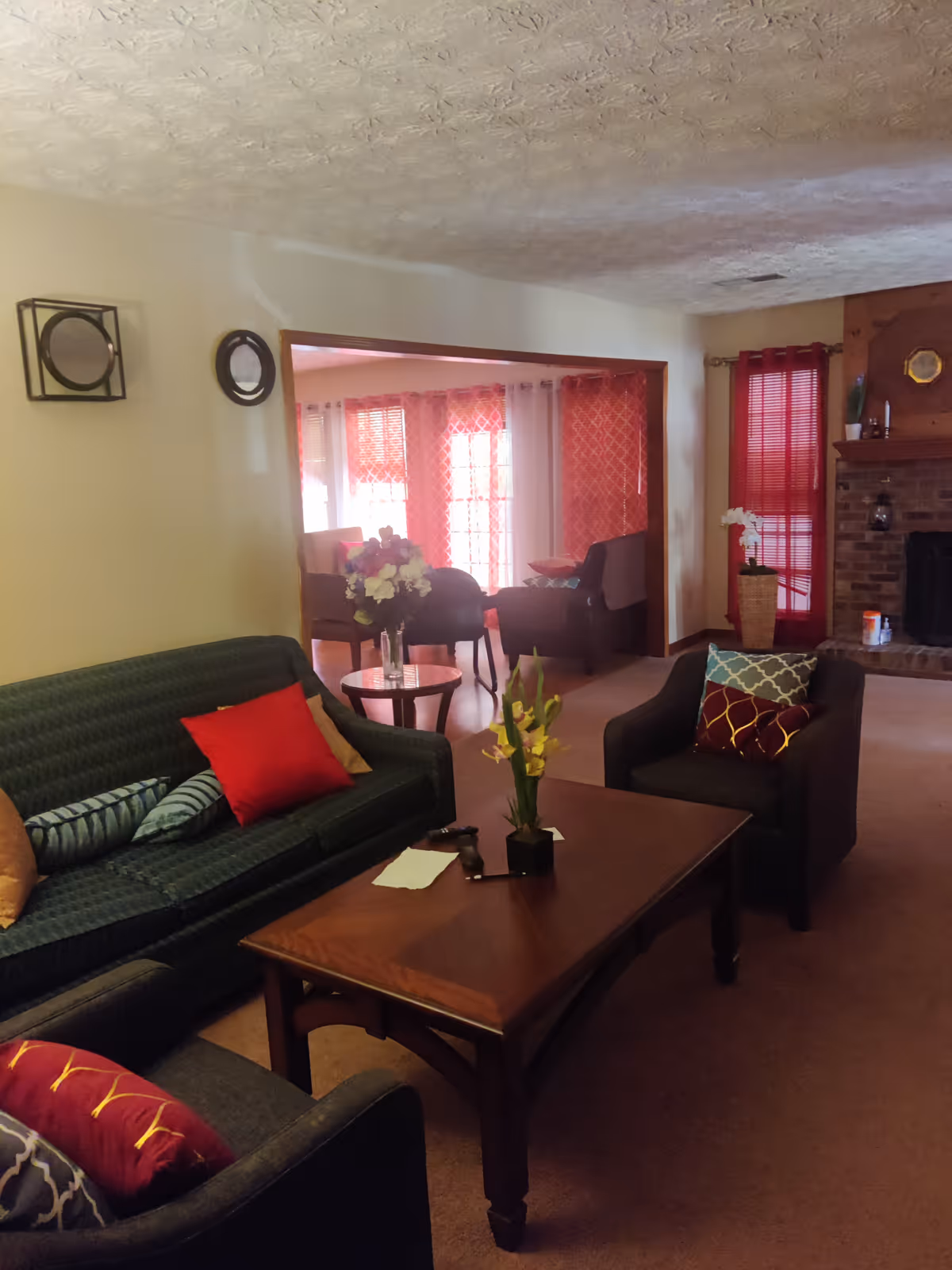 A cozy living room with a dark green sofa and two dark armchairs, each adorned with colorful cushions. A wooden coffee table with a small flower arrangement and some papers sits in the center. In the background, there is an adjoining room with large windows covered by red and white curtains, and a brick fireplace with a wooden mantle on the right side.