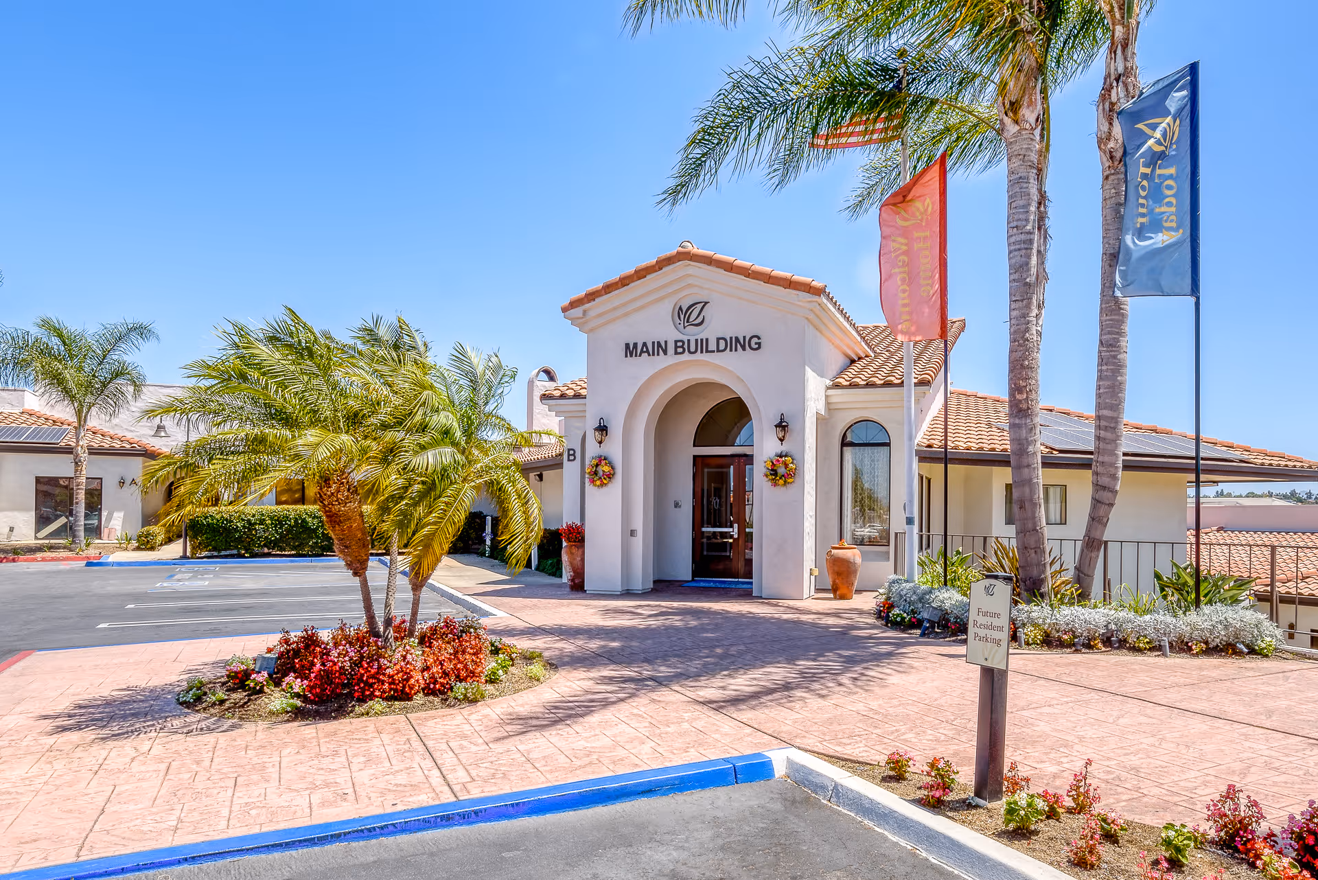 Front entrance of a senior living facility main building with palm trees, flags, and landscaped driveway.