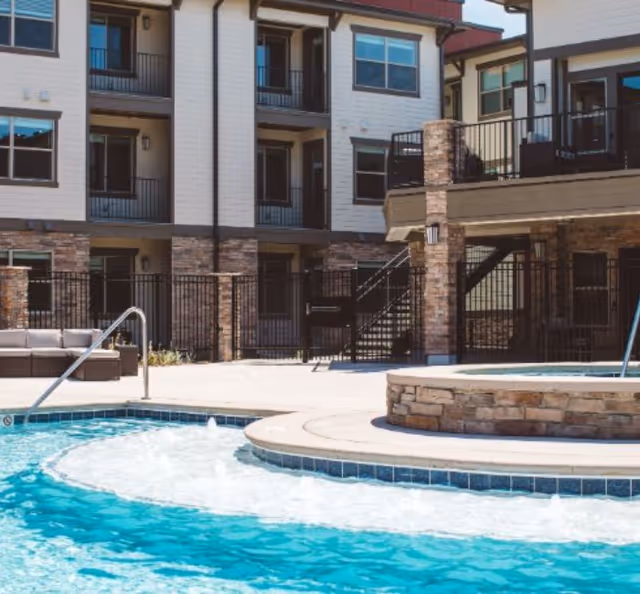 Resort-style outdoor pool and raised spa beside a multi-story residential building with balconies and patio seating.