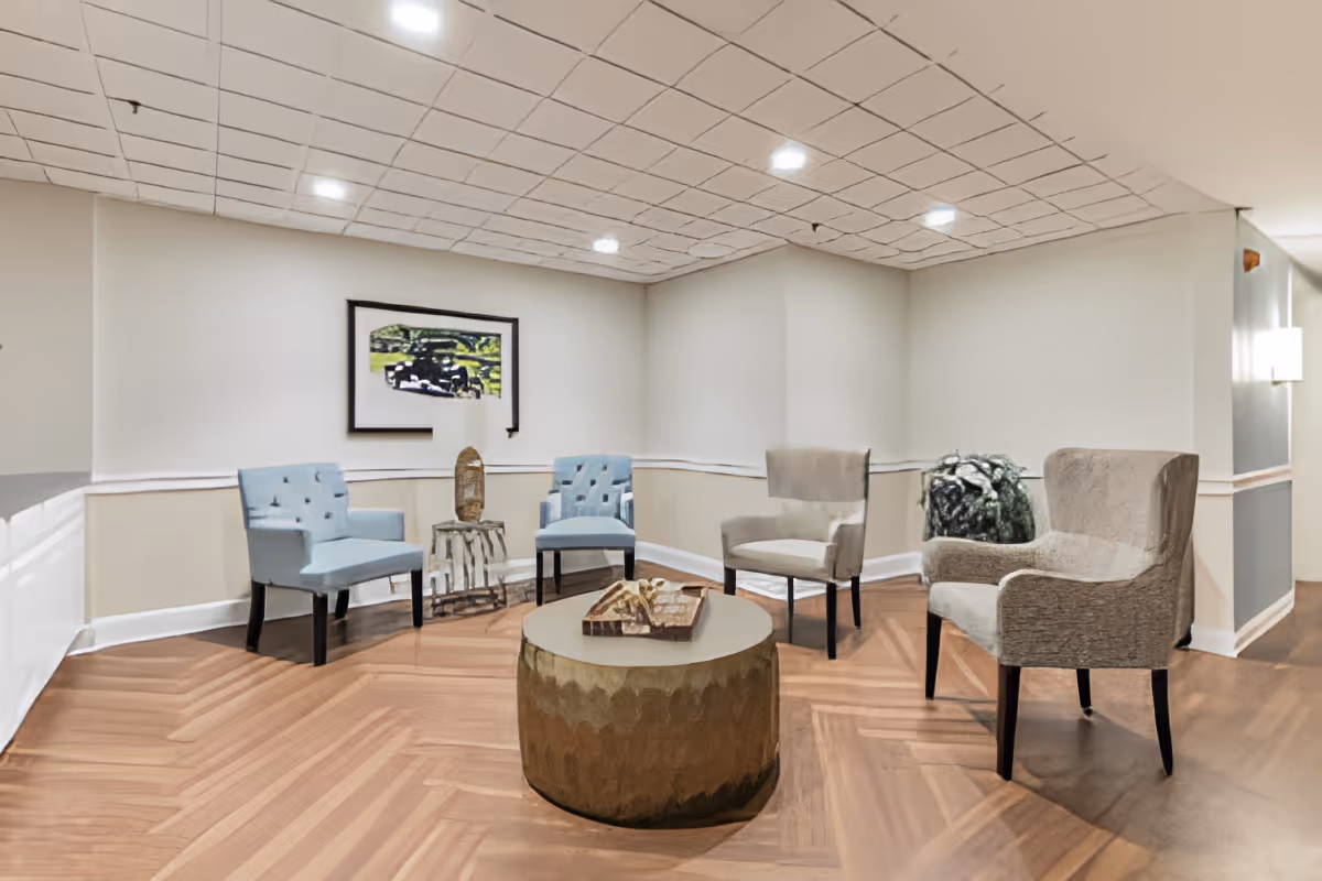 A cozy seating area in a senior living facility with four upholstered chairs arranged around a round coffee table. The room has wood-patterned flooring, white walls with beige wainscoting, a framed picture on the wall, and a decorative plant in the corner.