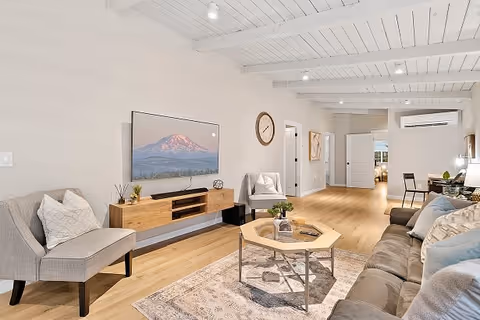 Bright open living room with a sofa, armchairs, coffee table, wall-mounted TV, and exposed white wood-beamed ceiling.
