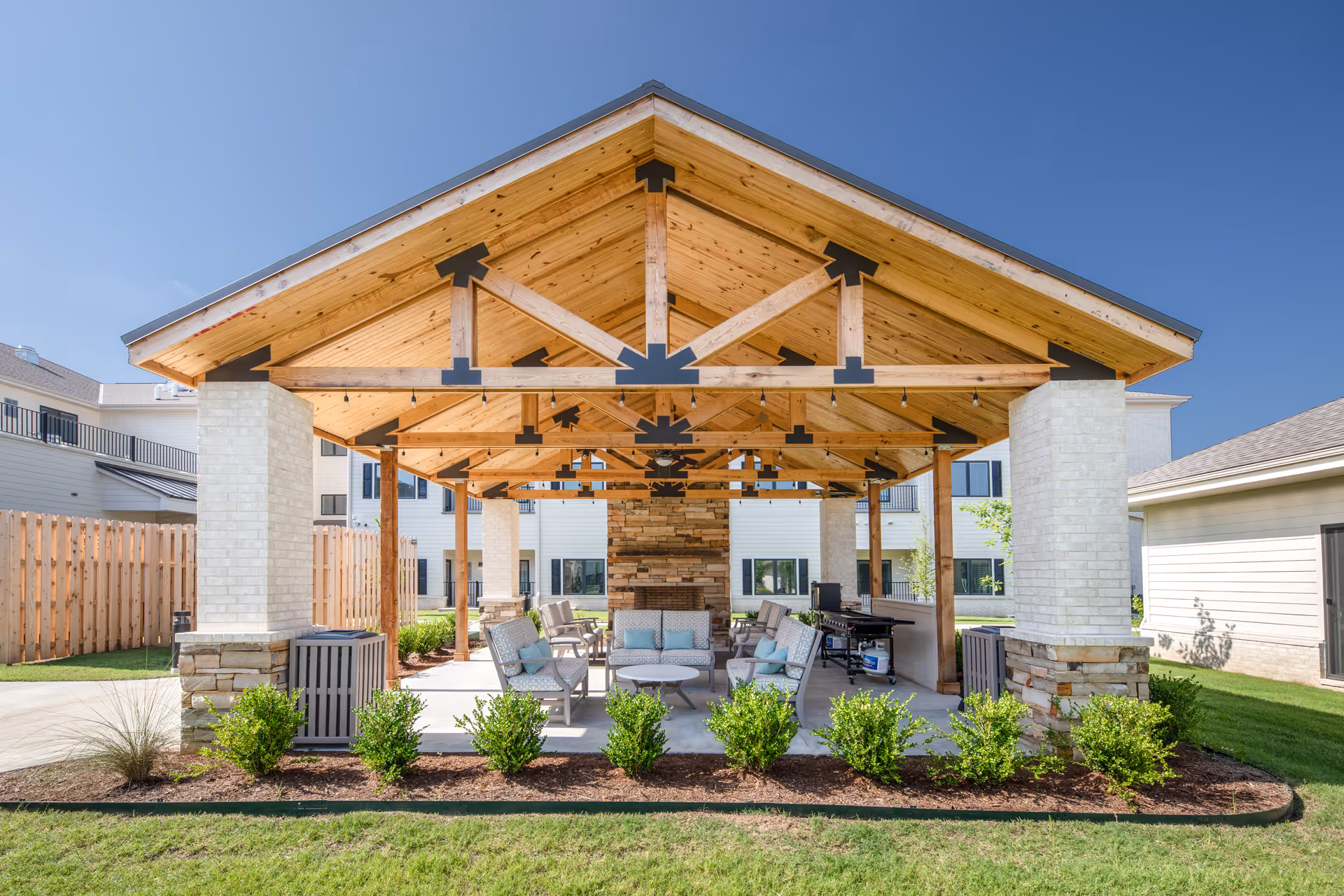 Outdoor covered seating area with wooden beams and a stone fireplace at the back, surrounded by chairs and small tables, with green bushes and grass in the foreground and residential buildings in the background under a clear blue sky.