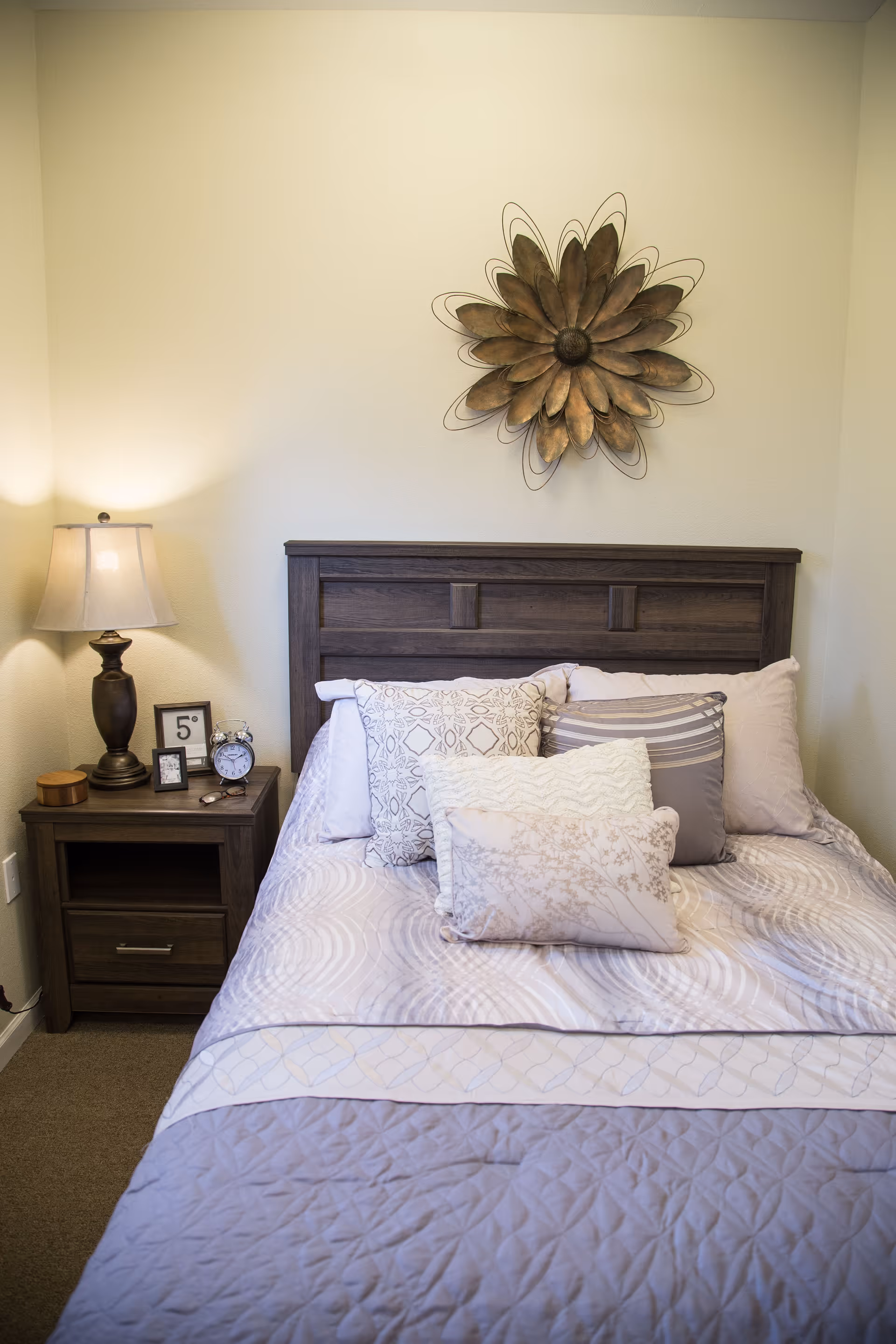 A neatly made bed with multiple decorative pillows in a bedroom. The bed has a wooden headboard and is covered with a light-colored quilt and patterned bedding. To the left of the bed is a wooden nightstand with a lamp, an alarm clock, a small framed picture, and a small wooden box. Above the bed on the wall is a large decorative metal flower art piece.