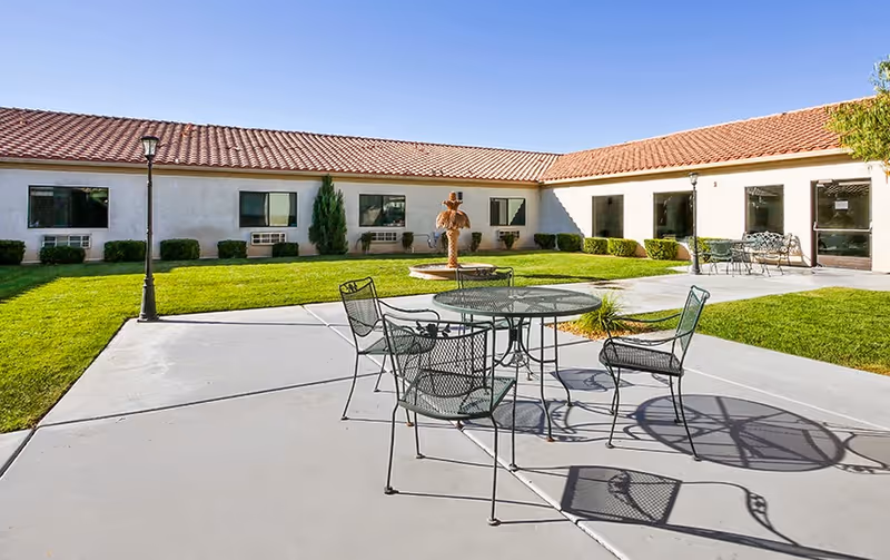 Outdoor courtyard area with metal patio table and chairs on a concrete surface, surrounded by green grass and a building with a red tile roof in the background. There is a small statue in the center of the grass and lamp posts near the seating area.
