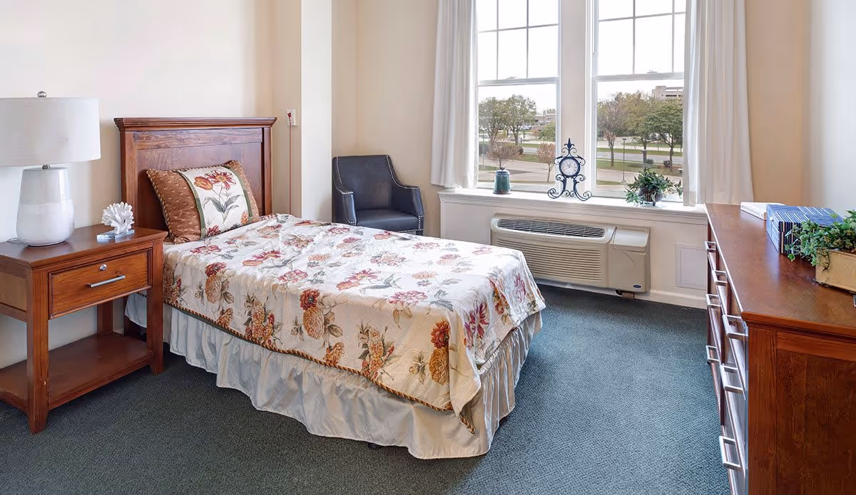 A cozy bedroom in a senior living facility featuring a single bed with floral bedding and a matching pillow. Next to the bed is a wooden nightstand with a white lamp and decorative coral piece. A black armchair is placed near a large window with white curtains, overlooking trees and a street outside. A wooden dresser with books and a plant is on the right side of the room. The room has a green carpet and beige walls.