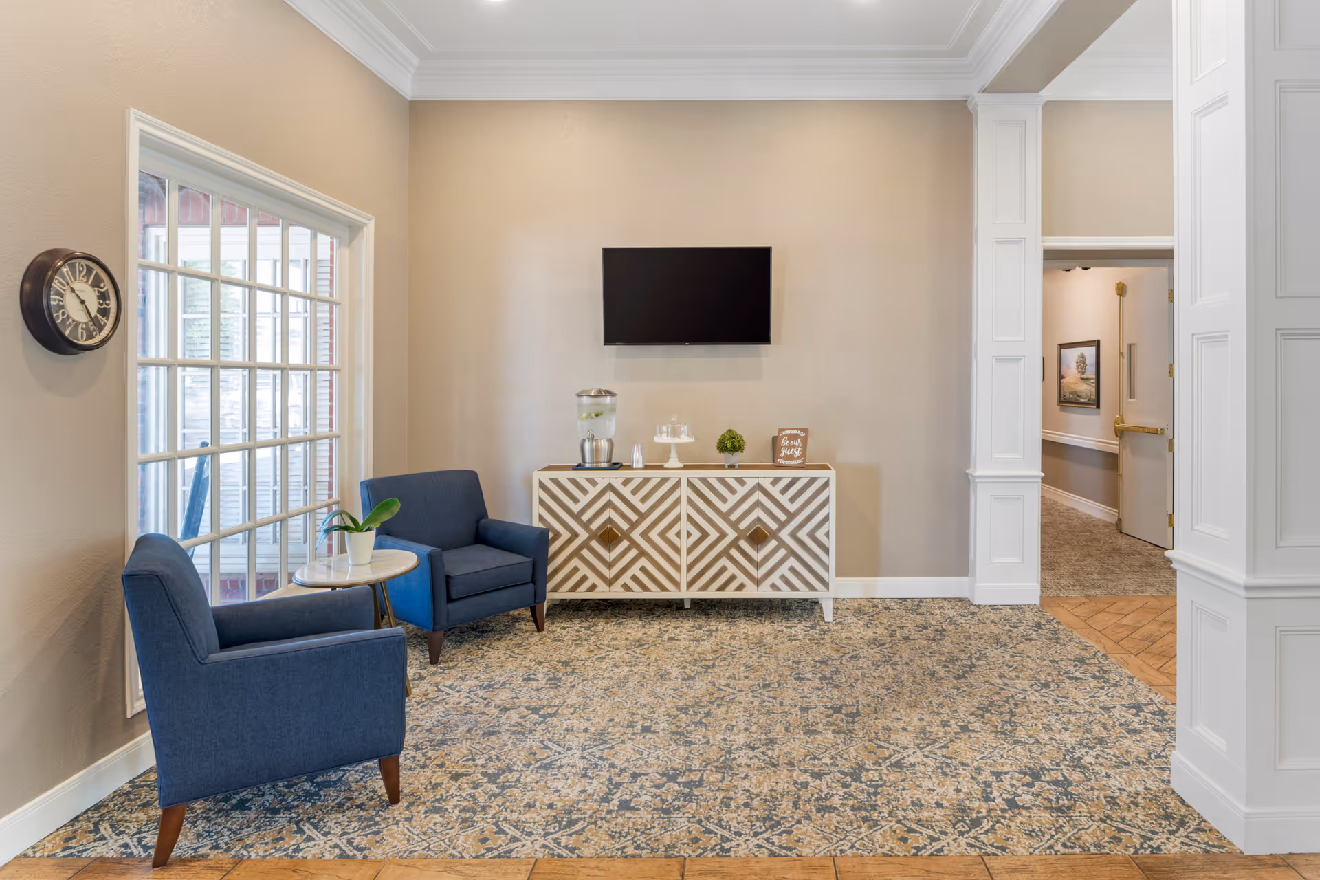 A cozy sitting area in a senior living facility with two blue armchairs around a small round table with a plant on it. A patterned cabinet is against the wall with a water dispenser, glasses, and a small plant on top. A flat-screen TV is mounted on the wall above the cabinet. There is a large window with multiple panes on the left side and a hallway entrance on the right.