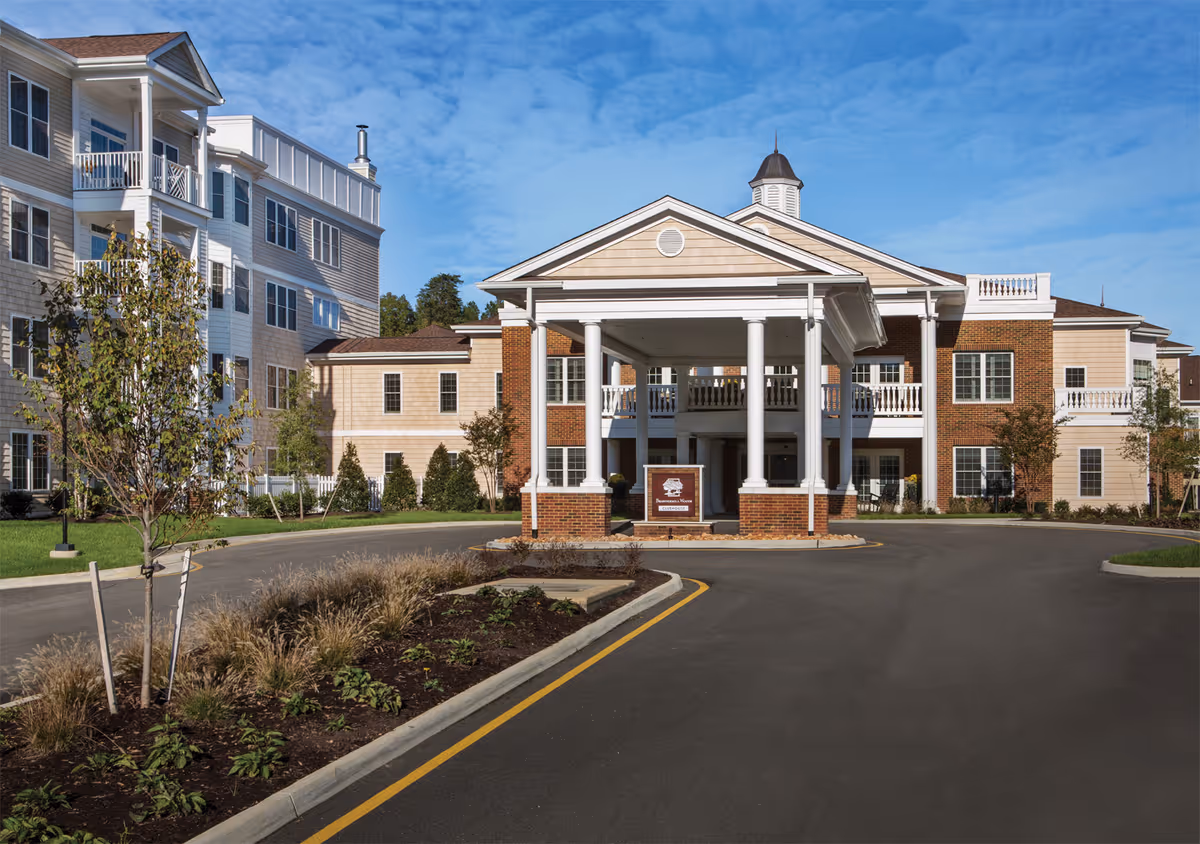 Front exterior view of Brandermill Woods Retirement facility showing a large building with beige and brick facade, white columns supporting a covered entrance, landscaped greenery, and a clear blue sky.