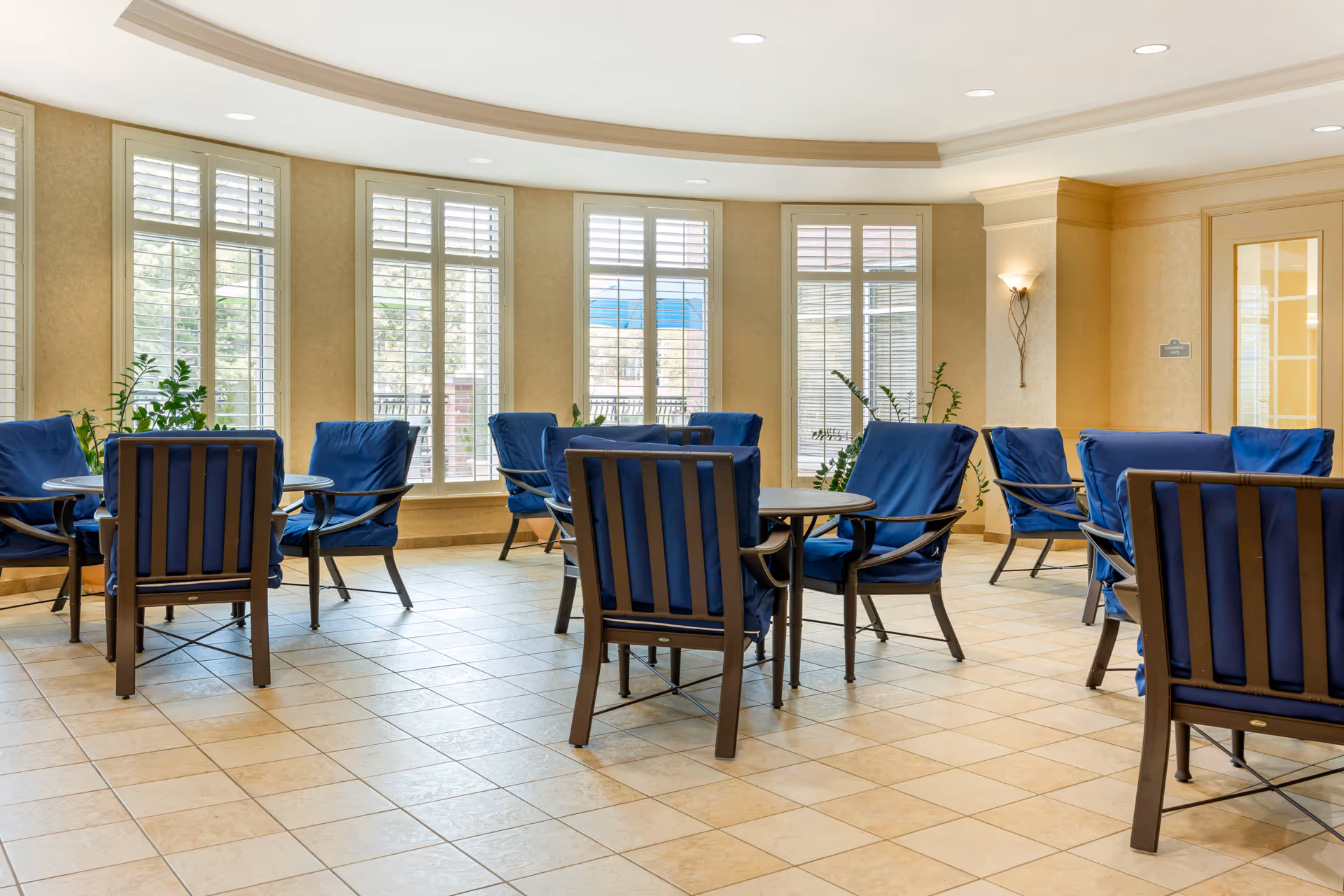 A bright and spacious common area with multiple round tables surrounded by blue cushioned chairs. Large windows with white shutters allow natural light to fill the room. There are some green plants near the windows and a wall sconce light fixture on the beige walls.