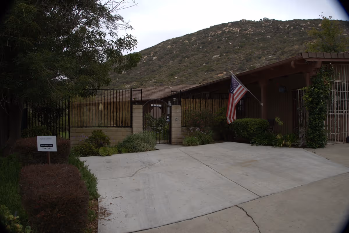 Entrance to Poway Gardens Senior Living facility with a gated fence, an American flag mounted on the building, and a sign indicating 'The Oaks'. There are bushes and plants around the entrance and a hill with rocks in the background.