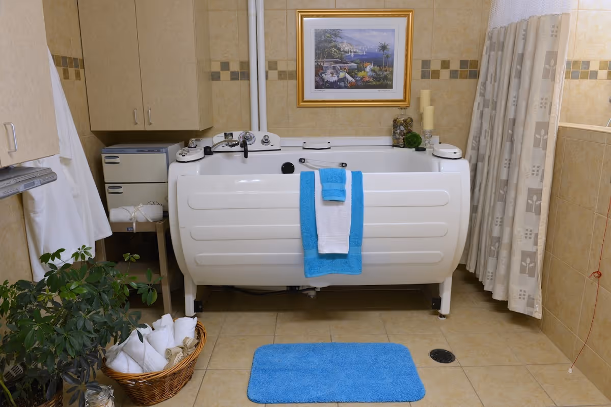 A tiled bathroom featuring a white walk-in therapeutic bathtub with blue and white towels, a blue bath mat, cabinets, plants and a basket of rolled towels.