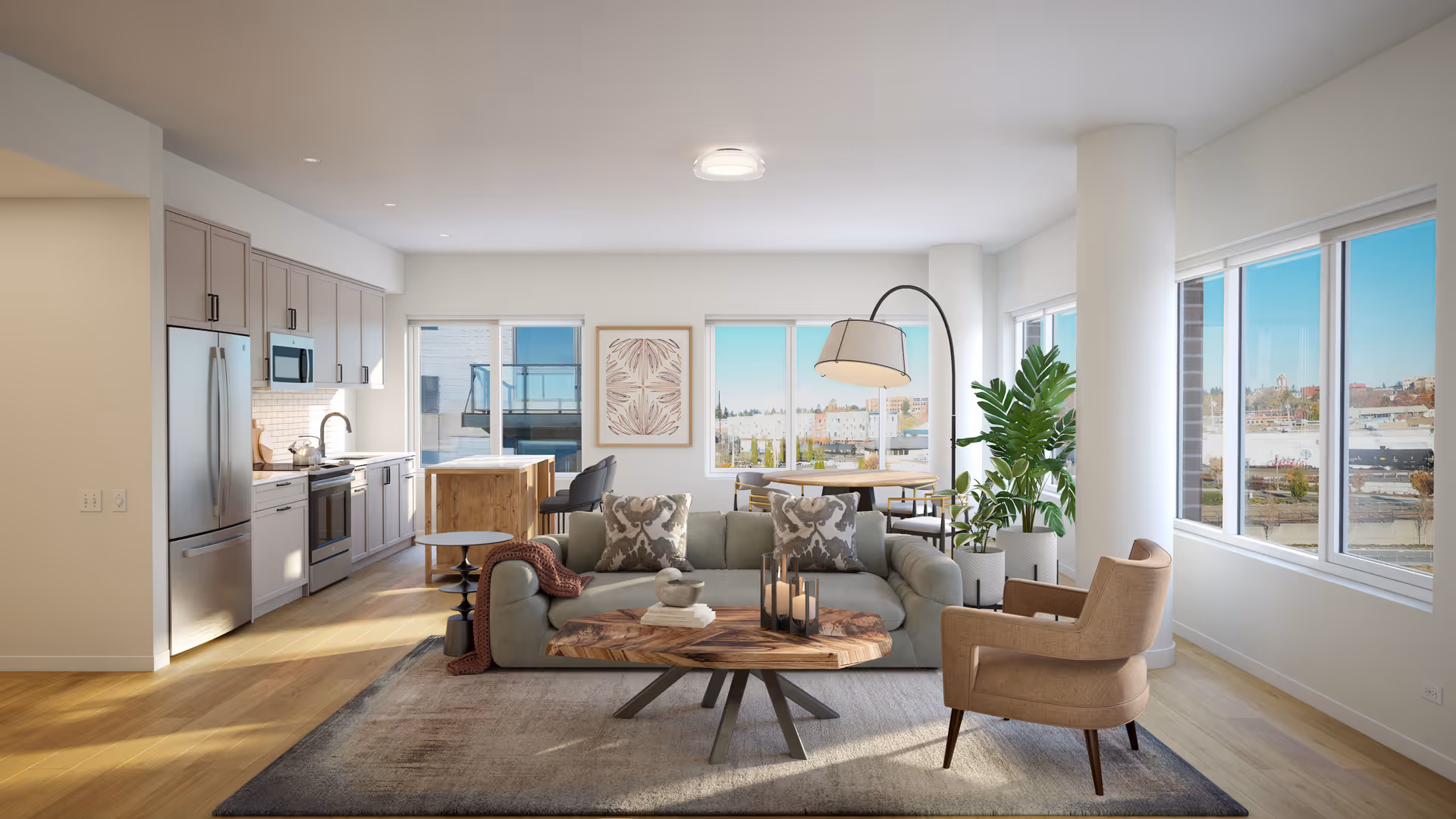 A bright and modern open-concept living space featuring a kitchen with stainless steel appliances and light gray cabinets on the left, a cozy living room area with a gray sofa, patterned pillows, a wooden coffee table, and a beige armchair in the center, and a dining area with a wooden table and chairs near large windows that offer a view of the city outside. The room has light wood flooring, white walls, and several large windows allowing natural light to fill the space.