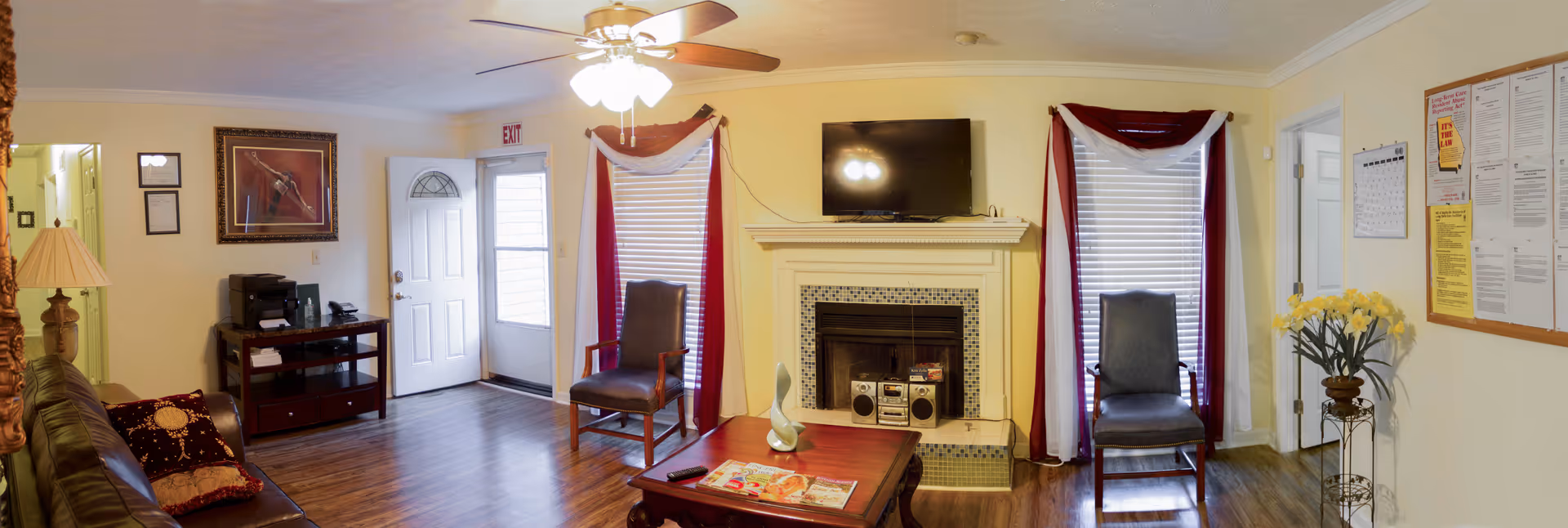 A cozy living room with a ceiling fan and light fixture, a white front door with a window, two windows with red and white curtains, a fireplace with a TV mounted above it, a wooden coffee table with magazines, two chairs, a brown leather couch with pillows, a wooden side table with a lamp, a printer on a small table, a bulletin board with notices on the wall, and a vase with yellow flowers on a stand.