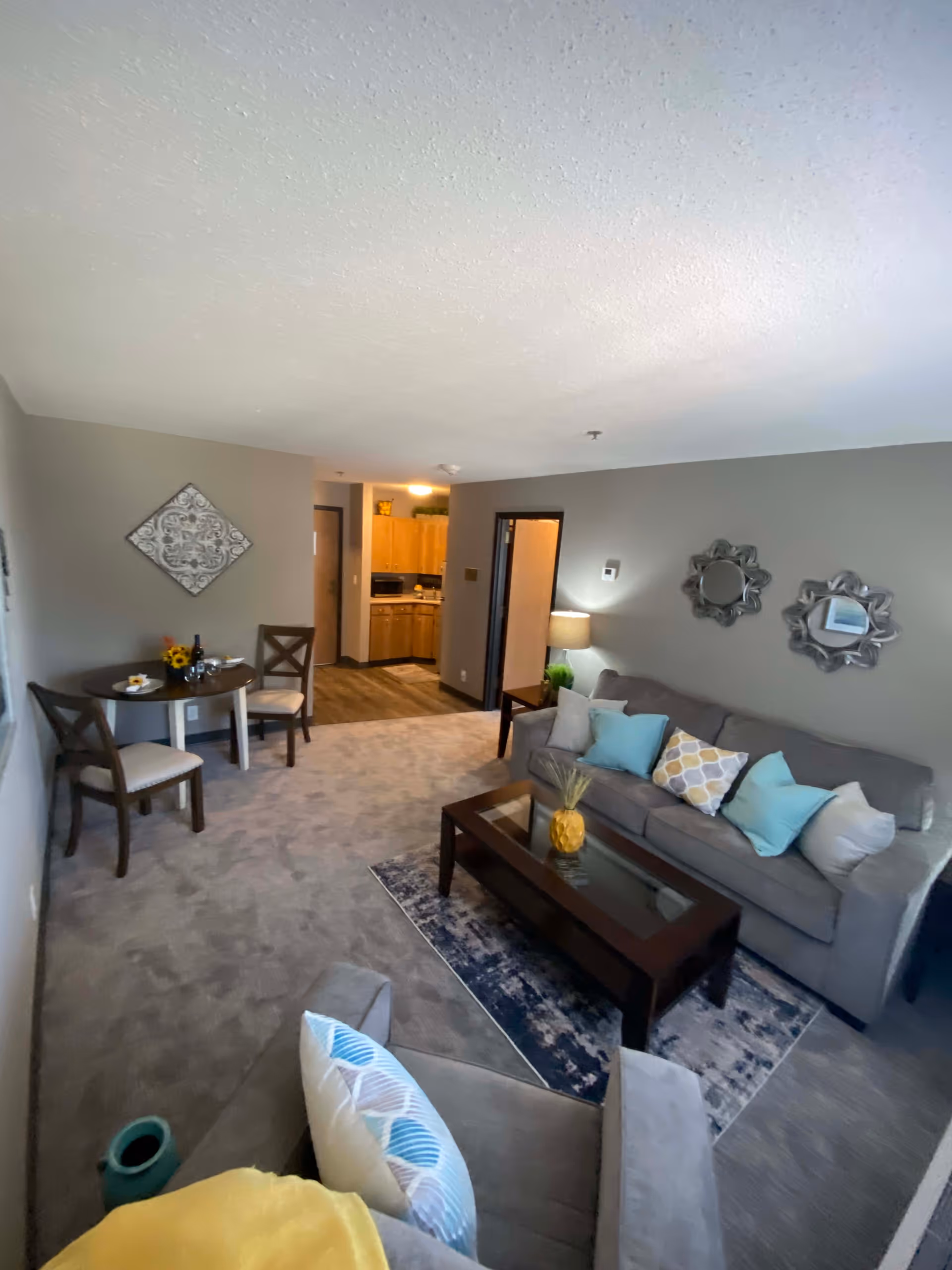 Interior view of a senior living facility's common area featuring a living room and dining space. The living room has a gray sofa with multiple decorative pillows, a glass-top coffee table with a pineapple decoration, and two decorative mirrors on the wall. Adjacent to the living room is a small round dining table with two chairs, set with a wine bottle, glasses, and a small flower arrangement. The kitchen area with wooden cabinets and appliances is visible in the background.