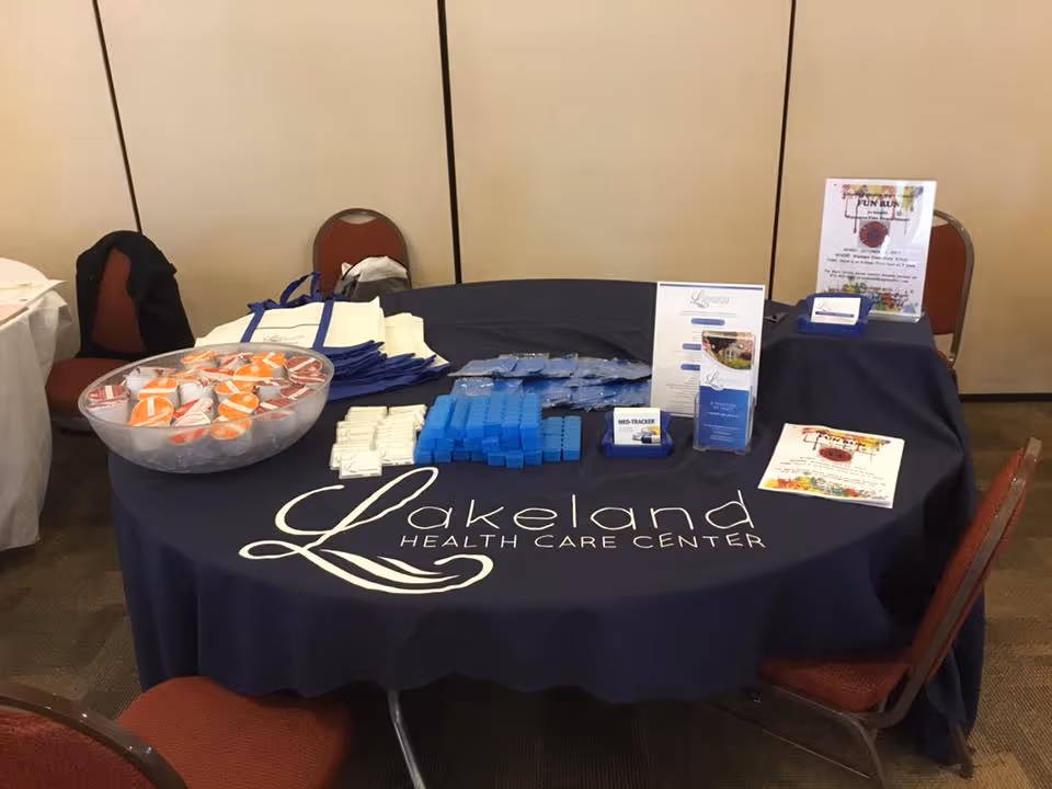 A round table covered with a dark blue tablecloth displaying the Lakeland Health Care Center logo. On the table are various promotional items including a bowl of orange and white cups, stacks of blue and white items, informational brochures, and flyers. The table is surrounded by red chairs and set against a beige partition wall.