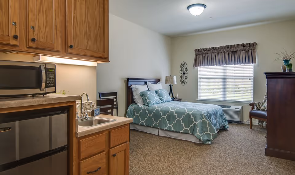 A senior living facility room featuring a bed with blue patterned bedding, a nightstand with a lamp, a window with blinds and a valance, a wooden chair, a wooden dresser, and a kitchenette area with a microwave, sink, and mini refrigerator.