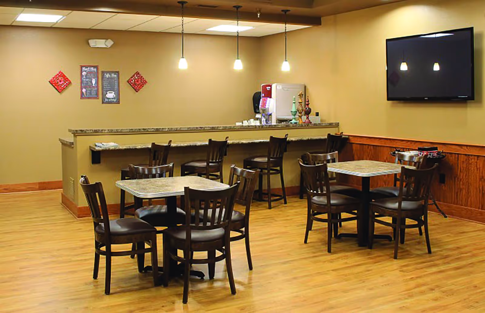 A dining area with two square tables, each surrounded by four wooden chairs with dark cushions. In the background, there is a counter with four high chairs and a coffee machine on the counter. The walls are beige with wooden trim, and a flat-screen TV is mounted on the right wall. Three pendant lights hang from the ceiling above the counter.