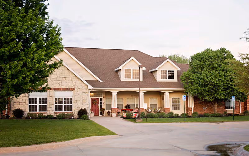 Exterior view of a single-story senior living facility building with a stone and brick facade, a brown shingled roof, and a covered entrance porch. There are two large trees on either side of the building, a well-maintained lawn, and a paved driveway with handicap parking signs visible.