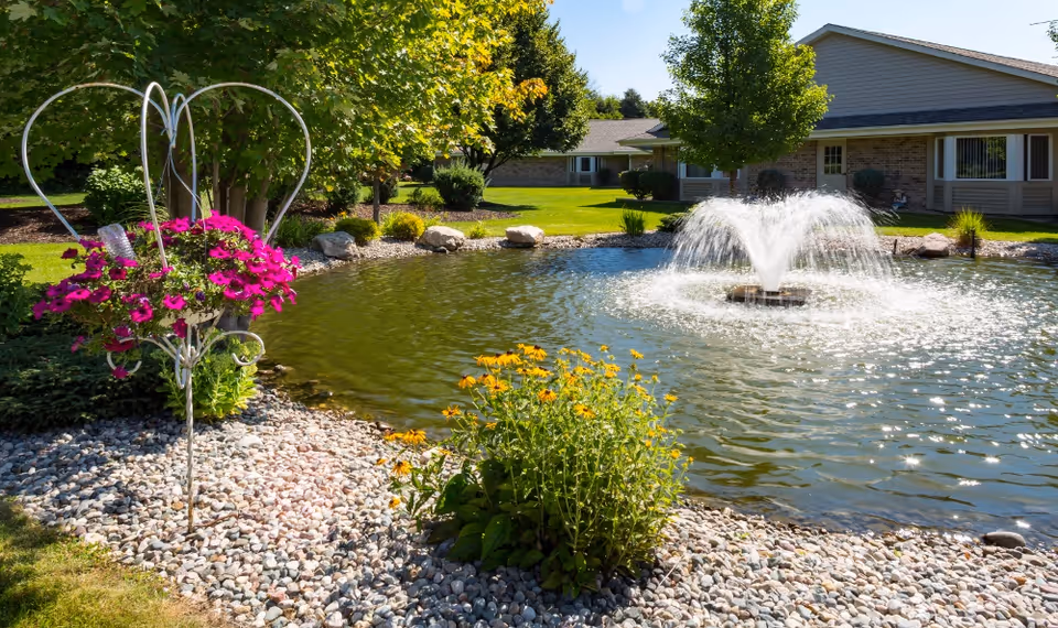 Landscaped pond with a central fountain, flowering planters, and single-story residential buildings in the background.