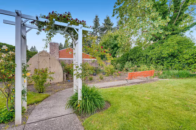 A garden area with a curved concrete pathway leading through a white wooden arbor adorned with climbing plants. There is a red bench on the right side of the path, surrounded by green grass, bushes, and trees. In the background, a small shed and a brick chimney are visible under a clear blue sky.