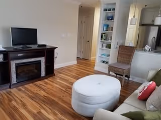 A cozy living room with wooden flooring, a TV on a dark wooden stand with an electric fireplace below, a white round ottoman in the center, a beige couch with colorful pillows, a wicker chair, and built-in shelves with decorative items. The room opens into a kitchen area with stainless steel appliances.