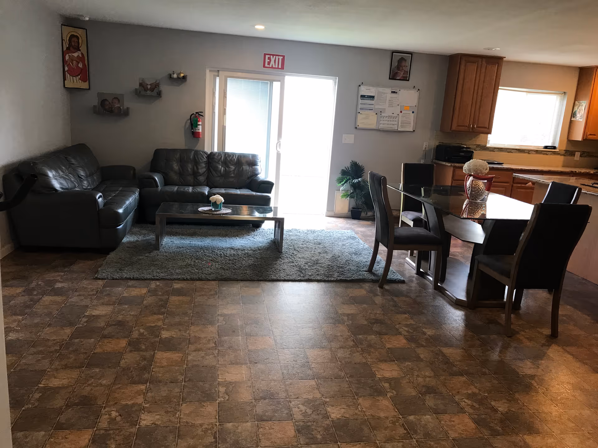 Interior view of a living and dining area in a senior care facility. The room features a gray leather sofa set with a glass coffee table on a gray rug. Adjacent to the seating area is a dining table with six chairs. The kitchen area with wooden cabinets and a window is visible in the background. There is an exit door with a red exit sign above it, a fire extinguisher on the wall, and some framed pictures and a bulletin board on the walls.