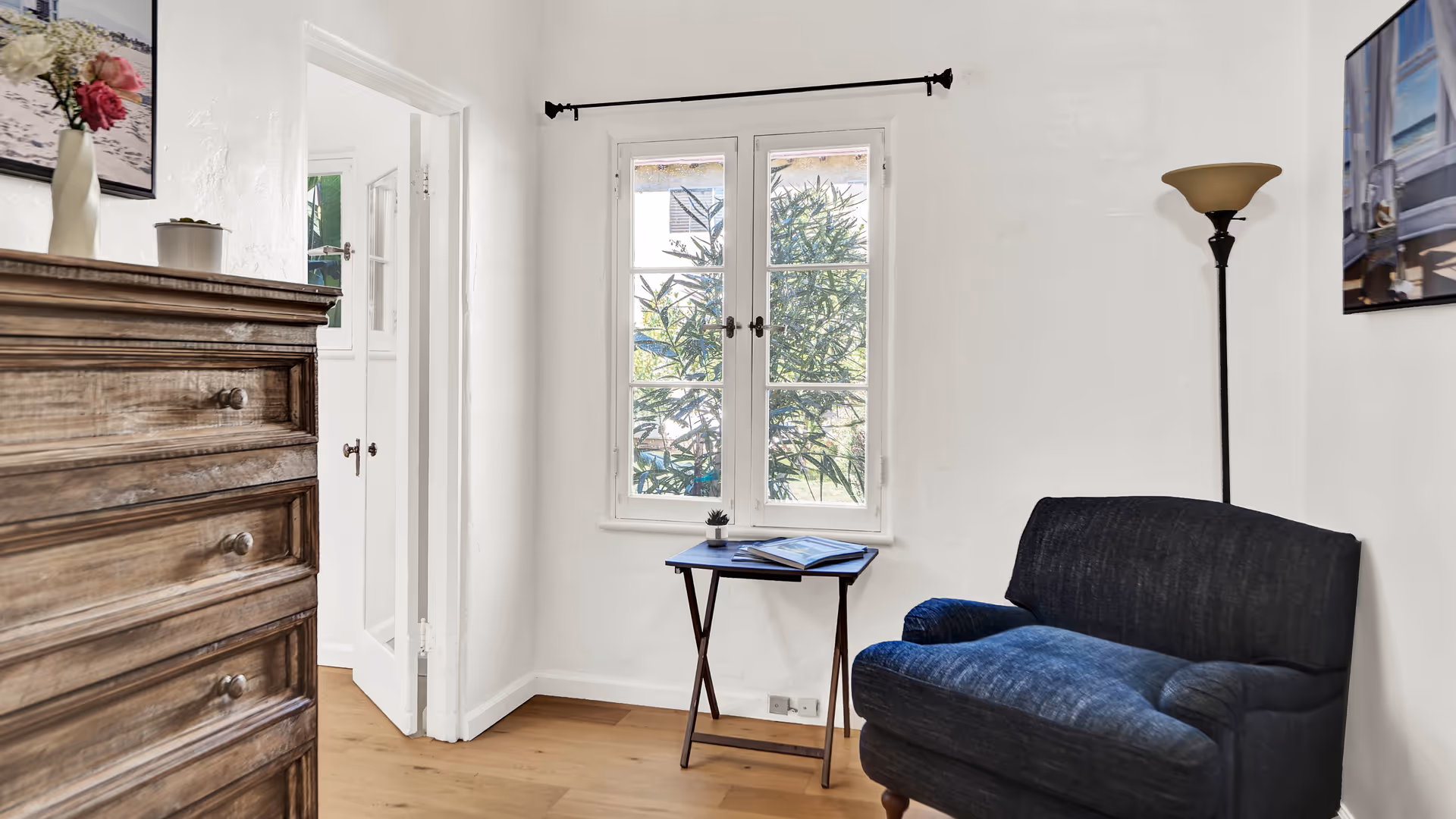 A cozy corner of a room with a dark blue armchair, a small wooden side table holding a small plant and an open book, a tall floor lamp with a beige shade, and a wooden chest of drawers with a vase of flowers on top. There is a window with white frames showing greenery outside and a white door leading to another room.