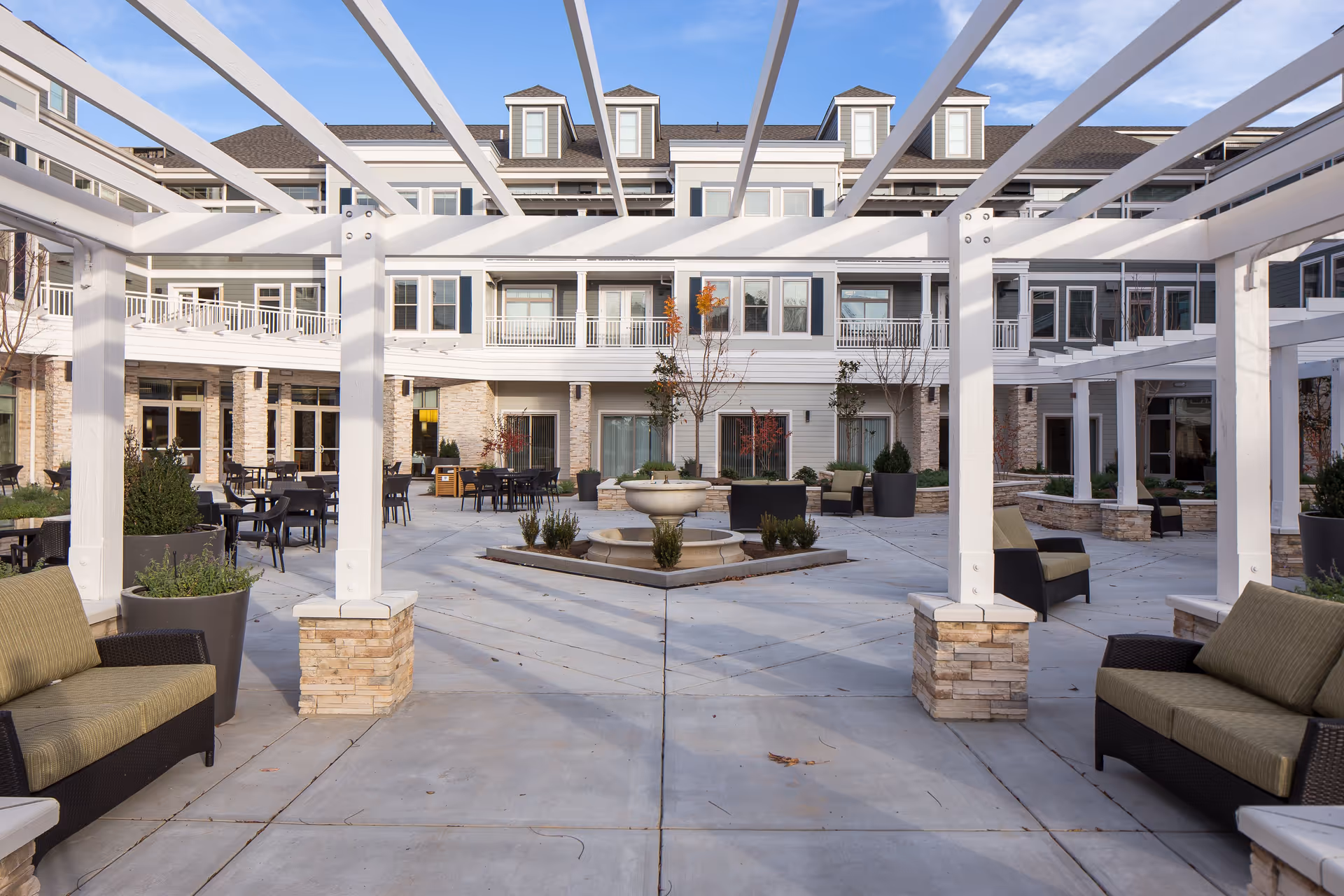Outdoor courtyard area at Merrill Gardens at Woodstock featuring white pergolas, cushioned seating, potted plants, tables and chairs, and a central water fountain with a multi-story building in the background under a clear blue sky.