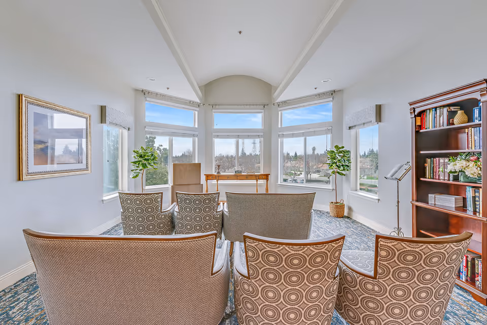 A bright room with large windows showing an outdoor view. The room has several chairs arranged in rows facing a wooden table at the front. There are two potted plants near the windows and a wooden bookshelf filled with books and decorative items on the right side. The walls are light-colored, and there is a framed picture on the left wall.