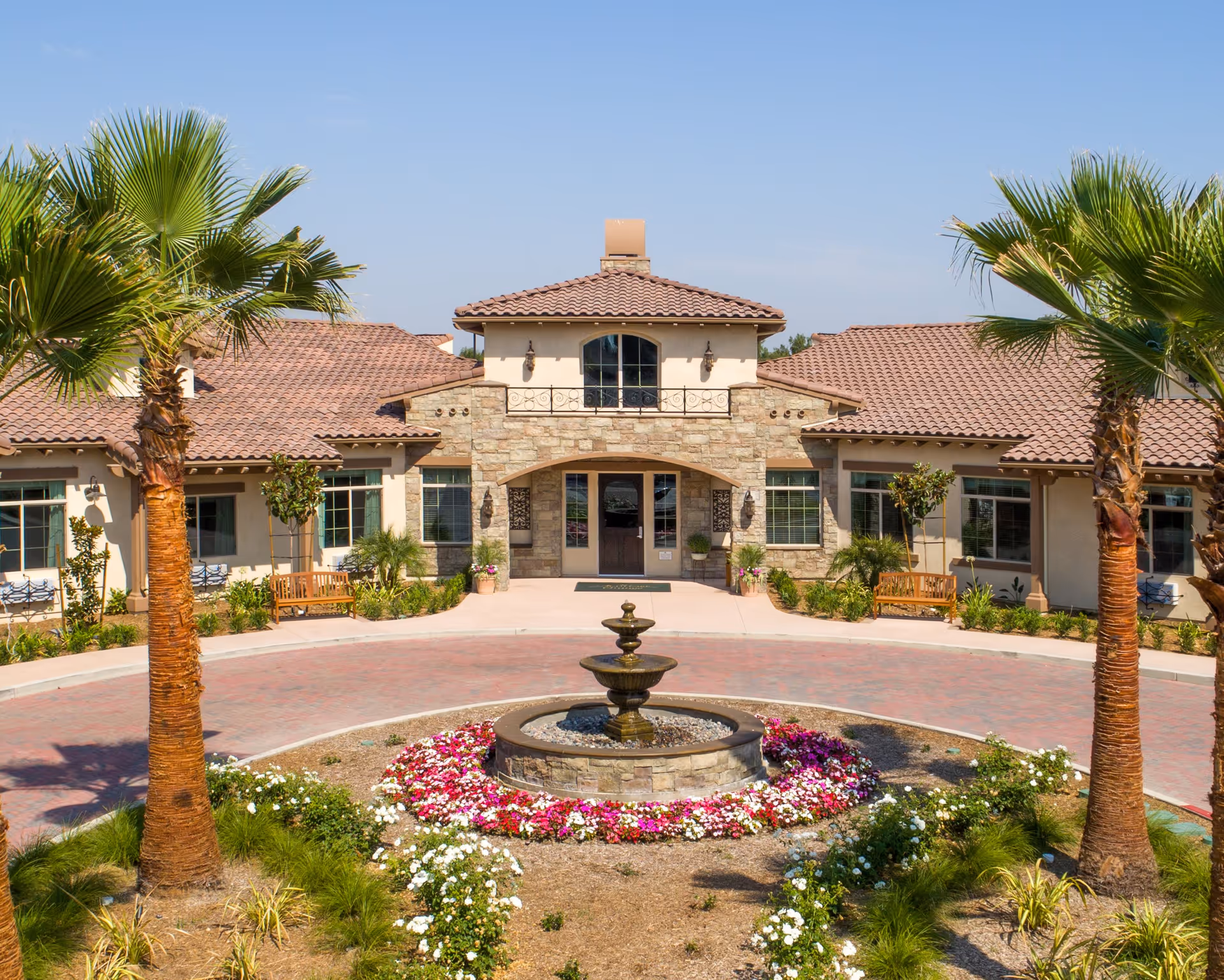 Front exterior view of a single-story building with a tiled roof, stone and stucco facade, surrounded by palm trees and landscaped flower beds with a central water fountain.