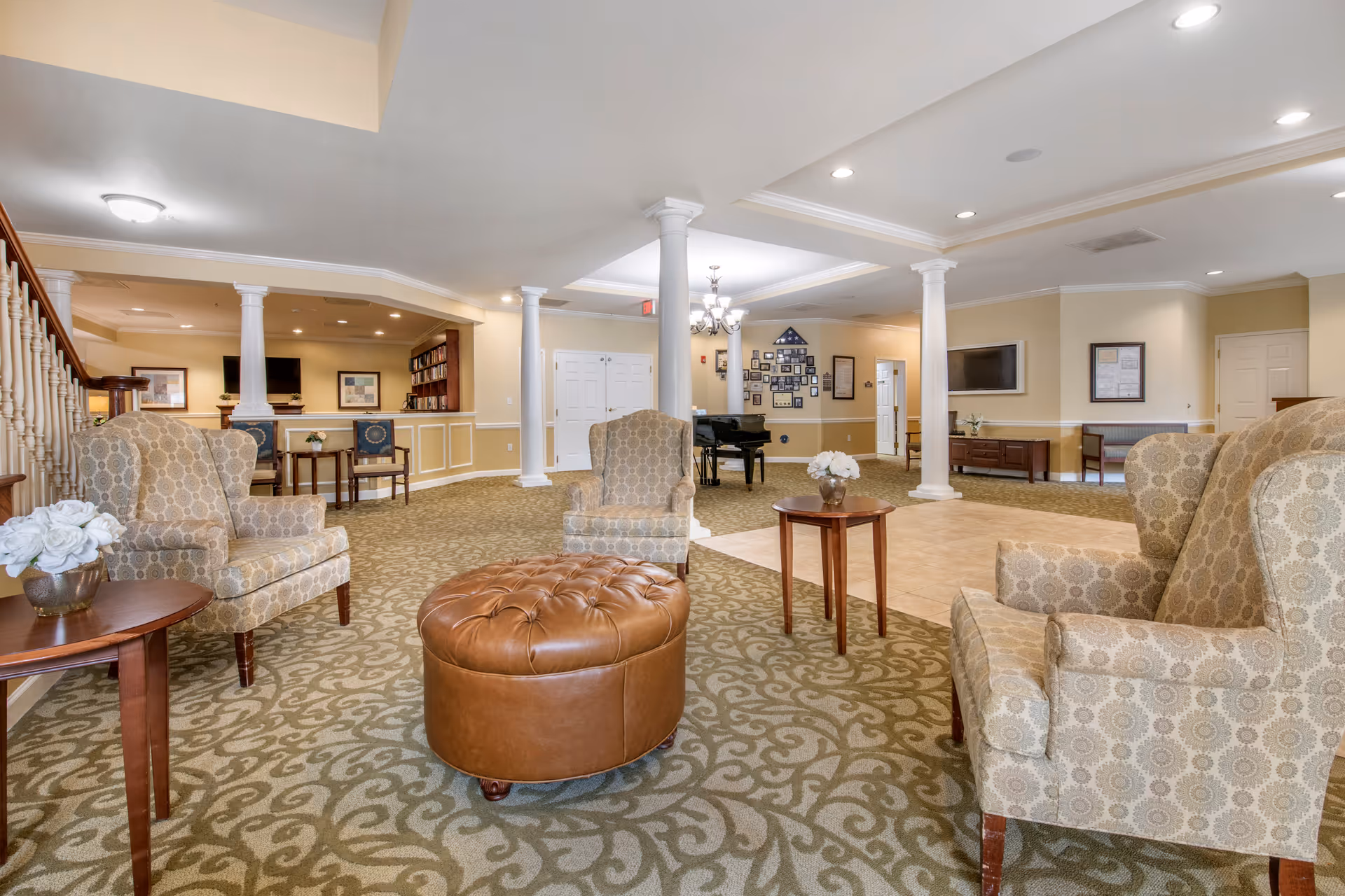 A spacious senior living facility common area with patterned carpet and beige walls. The room features several upholstered armchairs arranged around a round brown leather ottoman and small wooden tables with white floral arrangements. White columns support the ceiling, and there is a black grand piano in the background near a wall decorated with framed photos and an American flag display. Additional seating and a TV mounted on the wall are visible in the background.