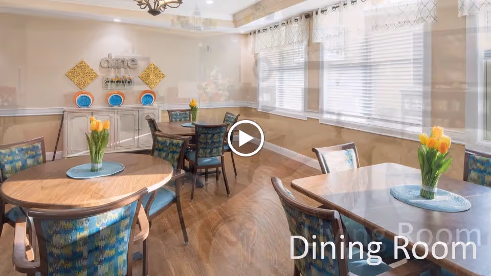 A bright dining room with several wooden tables and chairs featuring blue patterned upholstery. Each table has a vase with yellow tulips and a blue placemat. The room has large windows with white blinds and sheer curtains, wooden flooring, and a wall decoration with the word 'dine' above a sideboard.