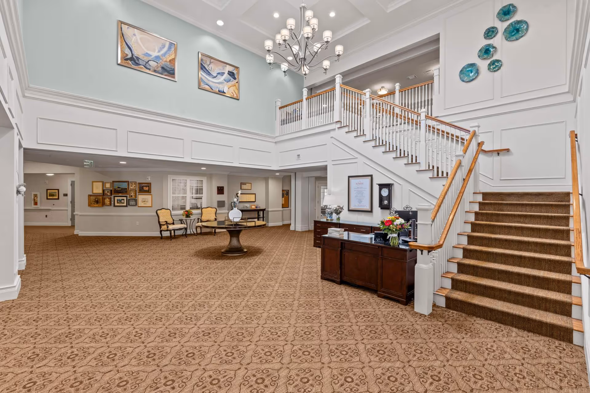 Bright two-story senior living lobby with a reception desk, carpeted staircase, seating area, chandelier, and wall art.