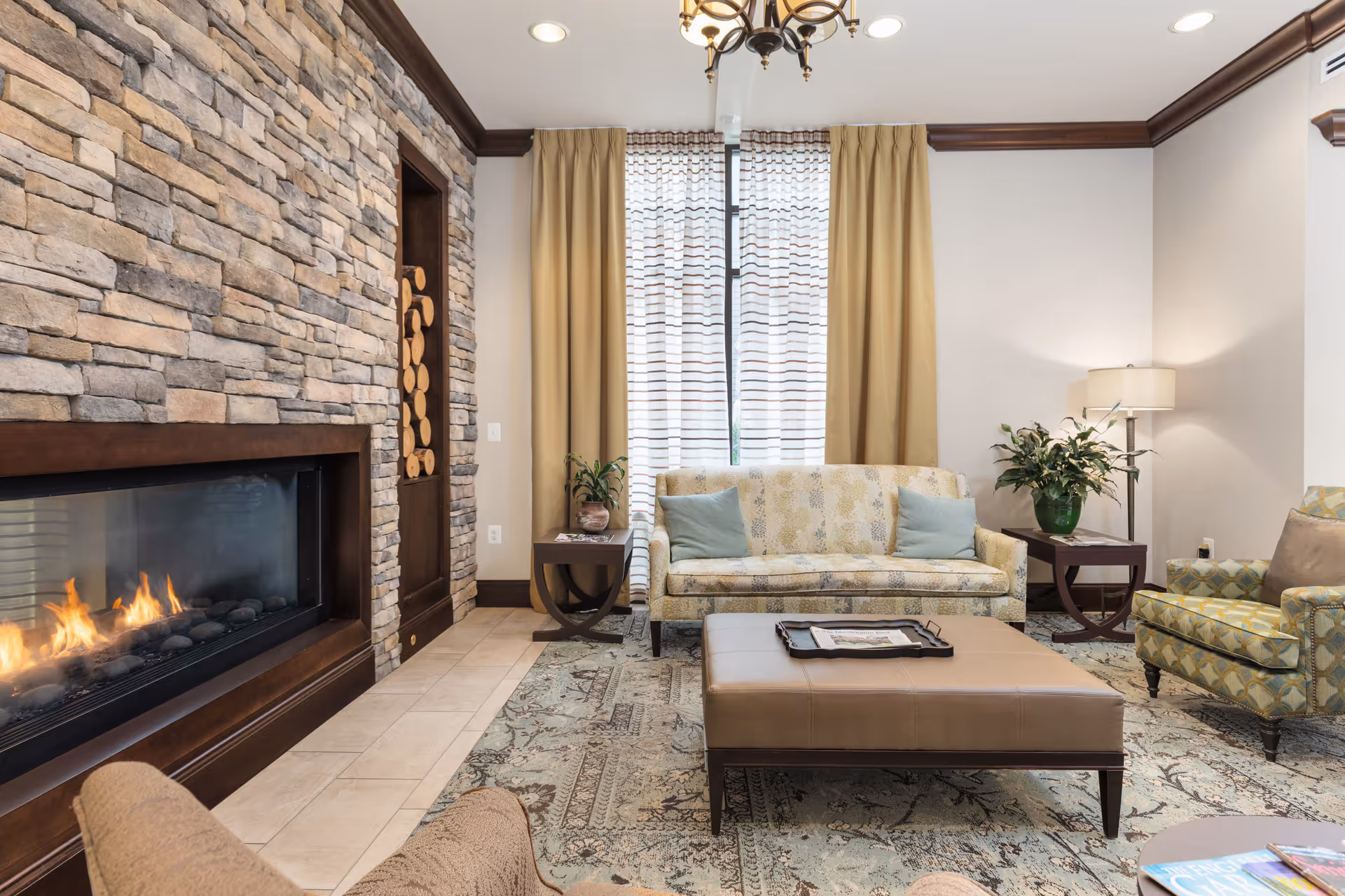 A cozy living room with a stone fireplace on the left side, a patterned sofa with two blue cushions in front of a window with beige curtains, a large ottoman coffee table in the center, an armchair with a geometric pattern on the right, side tables with plants and a lamp, and a patterned area rug covering the floor.