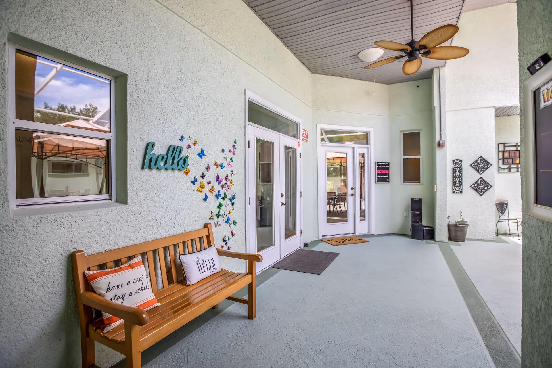 Covered entrance patio with a wooden bench, butterfly wall decor and double glass doors leading into the facility.