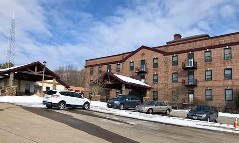 Exterior view of a multi-story brick building with several windows and small balconies. There are four cars parked in front of the building on a paved area with patches of snow. A covered entrance is visible, and a tall communication tower stands in the background under a partly cloudy sky.