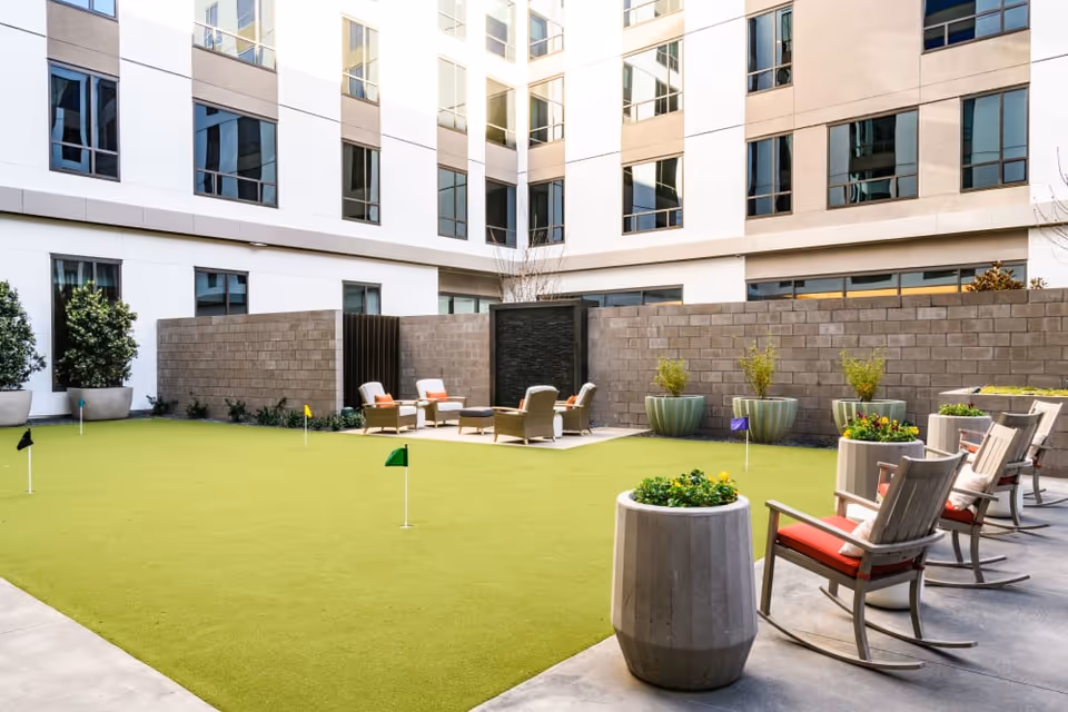 Outdoor courtyard with an artificial putting green, seating area with chairs and planters surrounded by a multi-story building.