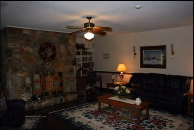 A cozy living room with a stone fireplace decorated with small house figurines. There is a ceiling fan with a light, a dark leather couch, a wooden coffee table with a floral centerpiece, a patterned area rug, a side table with a lamp, and a framed picture on the wall.