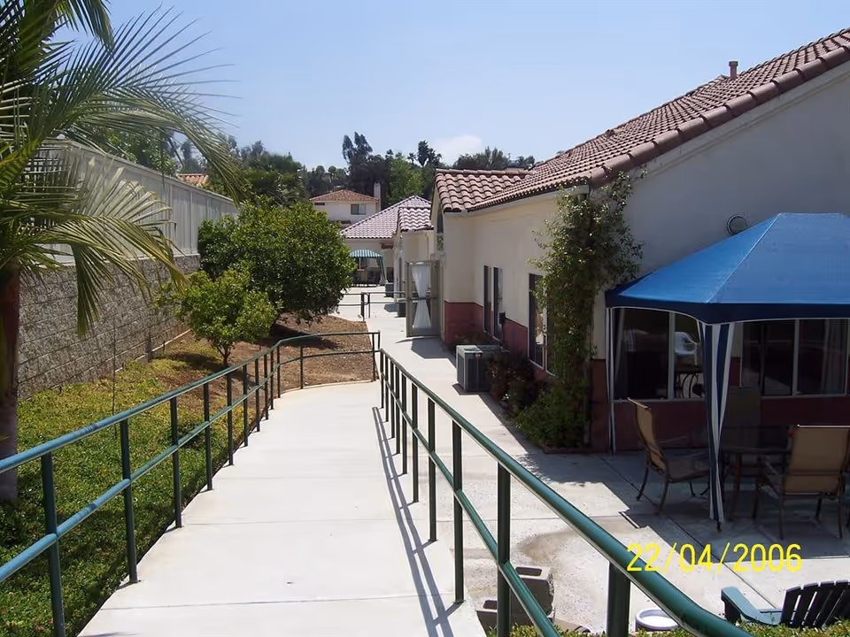Concrete ramp with green handrails leading past a single-story building with a patio canopy and landscaping.