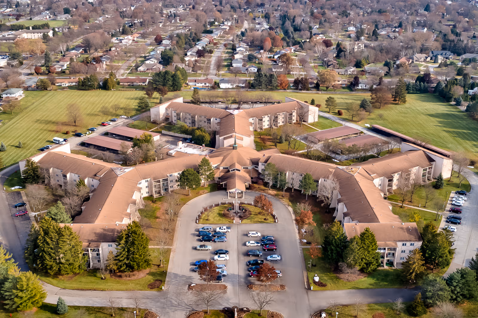 Aerial view of the Holiday Winter Village senior living complex showing interconnected three-wing buildings, a circular entrance drive with parked cars, and surrounding lawns and neighborhood.